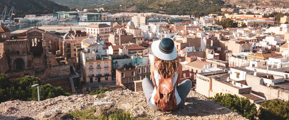 A woman sitting on a rocky outcrop overlooking a coastal town with historic buildings and mountains in the background.