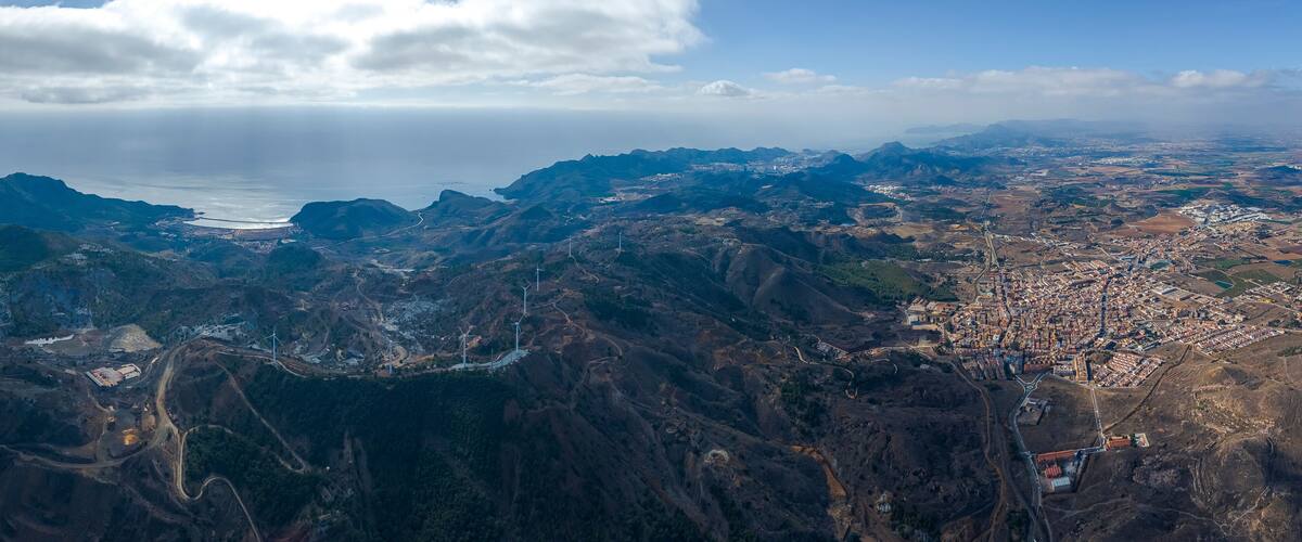 A mountainous landscape with a coastal city, wind turbines, and a winding road.