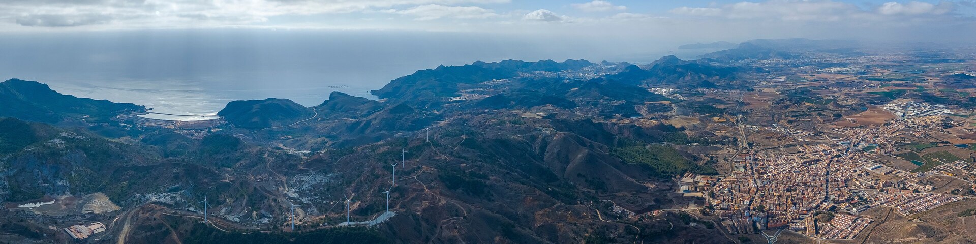 A mountainous landscape with a coastal city, wind turbines, and a winding road.