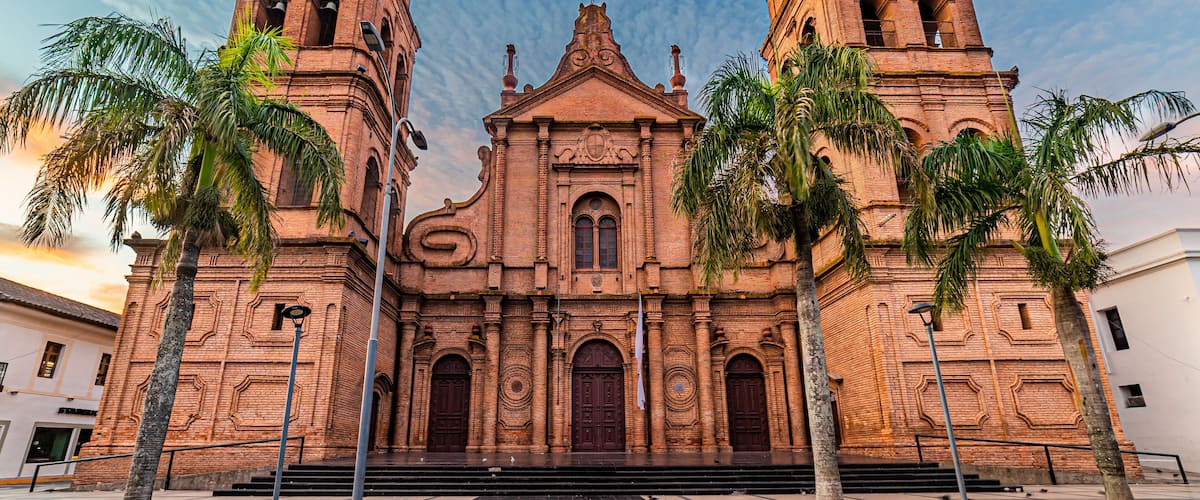 A large brick church with two bell towers and palm trees in front.