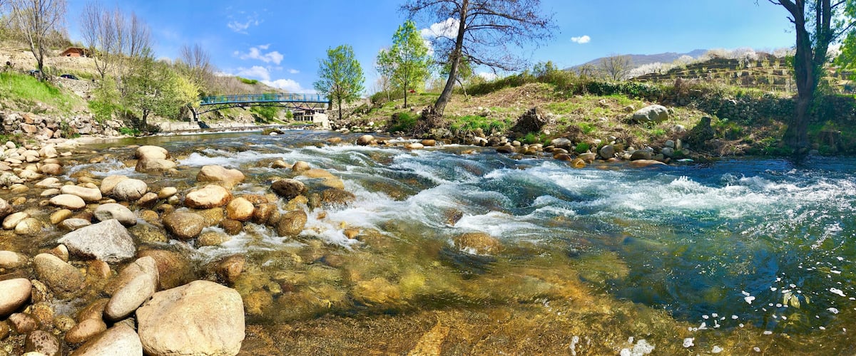 A river with clear water, rocks, a bridge, trees, and a hilly landscape.