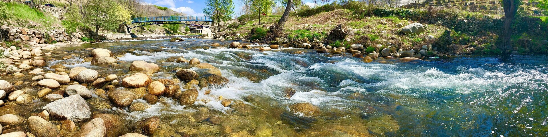 A river with clear water, rocks, a bridge, trees, and a hilly landscape.