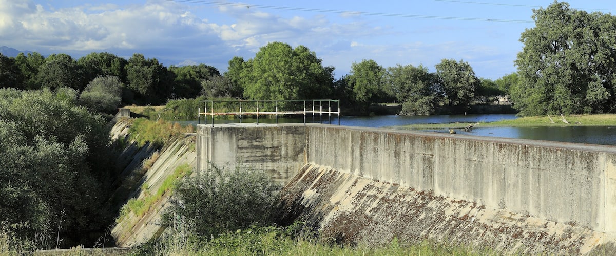 A concrete dam with a metal railing, a river, and surrounding greenery.