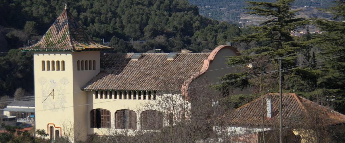 A historic building with a tower and arched windows, surrounded by trees and hills.