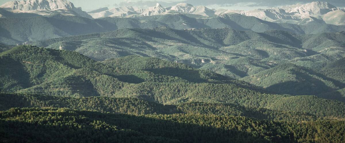 A mountainous landscape with dense forests and snow-capped peaks under a cloudy sky.