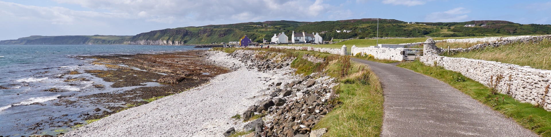 Un sendero costero con un muro de piedra, playa rocosa y casas al fondo.