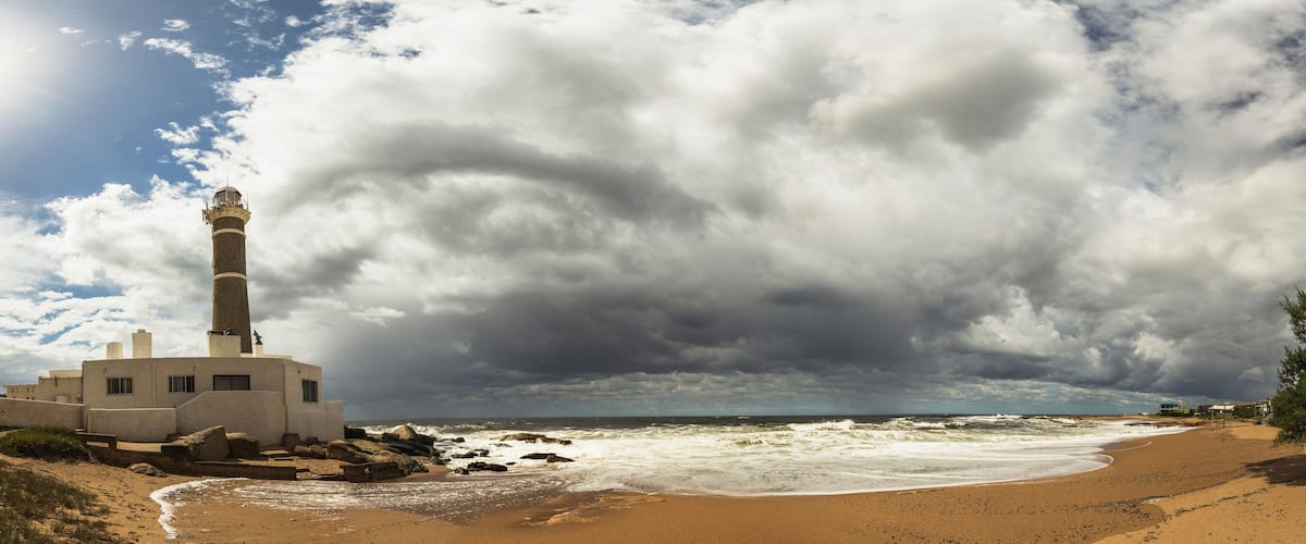 A coastal scene with a lighthouse, beach, rocky shoreline, and stormy sky.