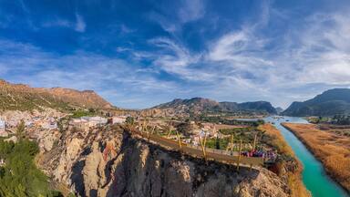 A suspension bridge over a river, a town below, and mountains in the background.