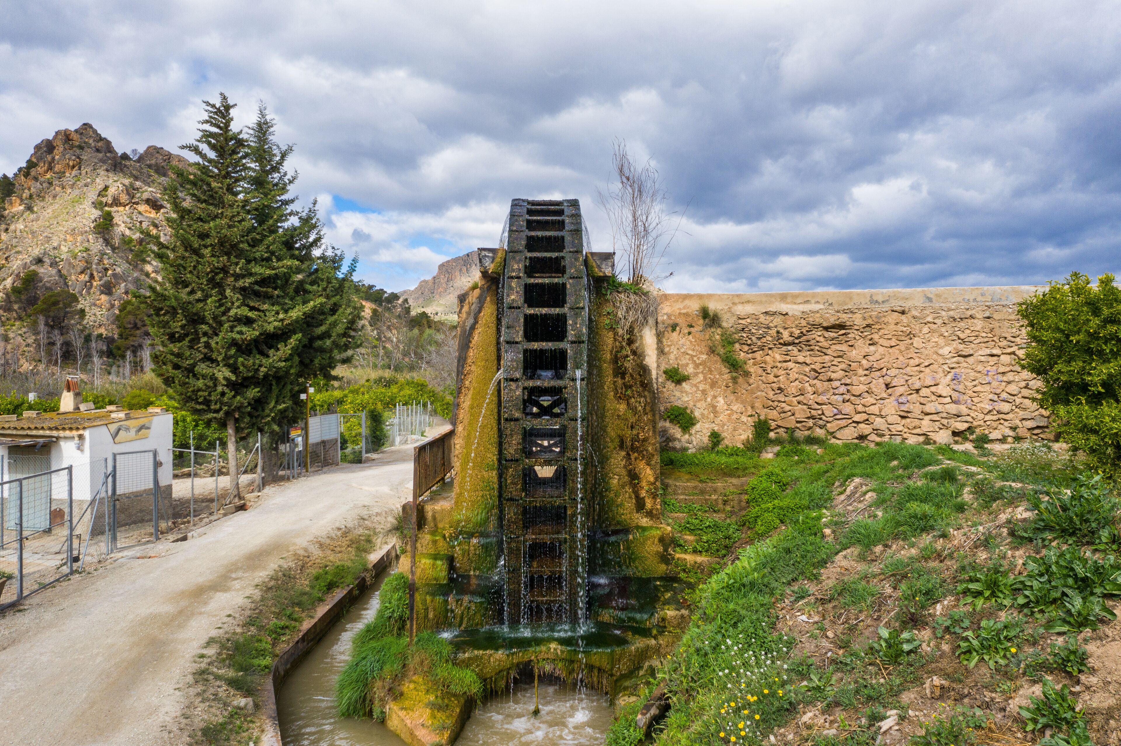 A stone water tower with a staircase leading to a water spout, surrounded by greenery and a rocky landscape.