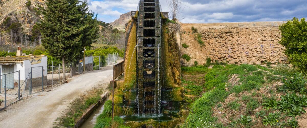 A stone water tower with a staircase leading to a water spout, surrounded by greenery and a rocky landscape.