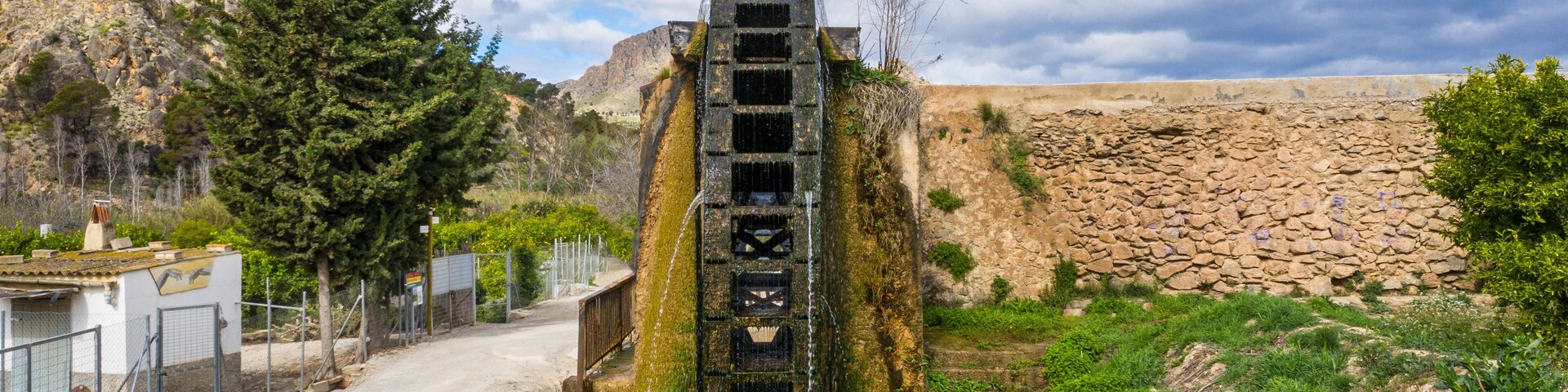 A stone water tower with a staircase leading to a water spout, surrounded by greenery and a rocky landscape.