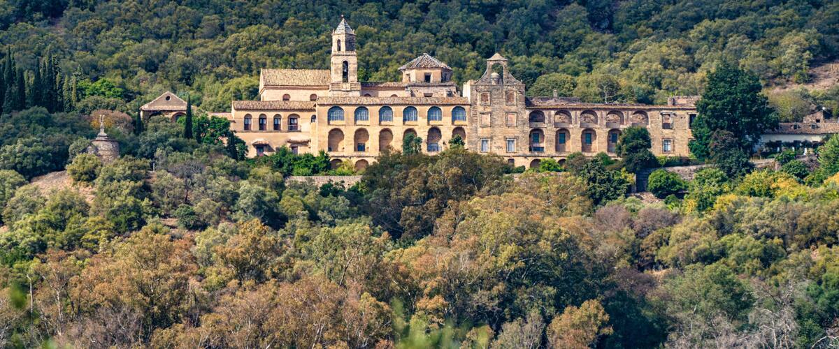 A historic building with multiple arches and a bell tower, surrounded by dense forest.