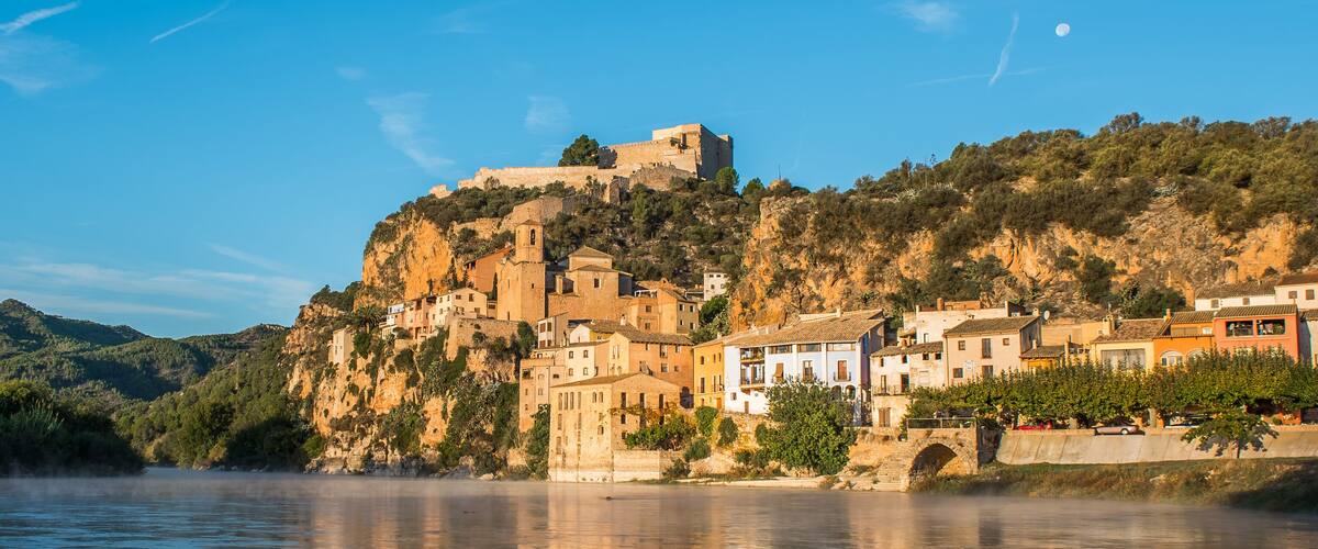 A coastal town with historic buildings on a hillside, a river in the foreground, and a clear sky.