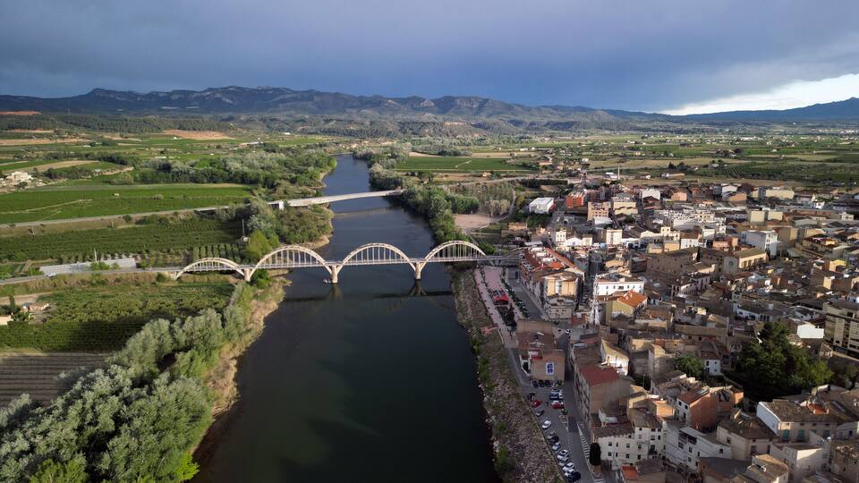 A river flowing through a town with a bridge, surrounded by mountains and fields.