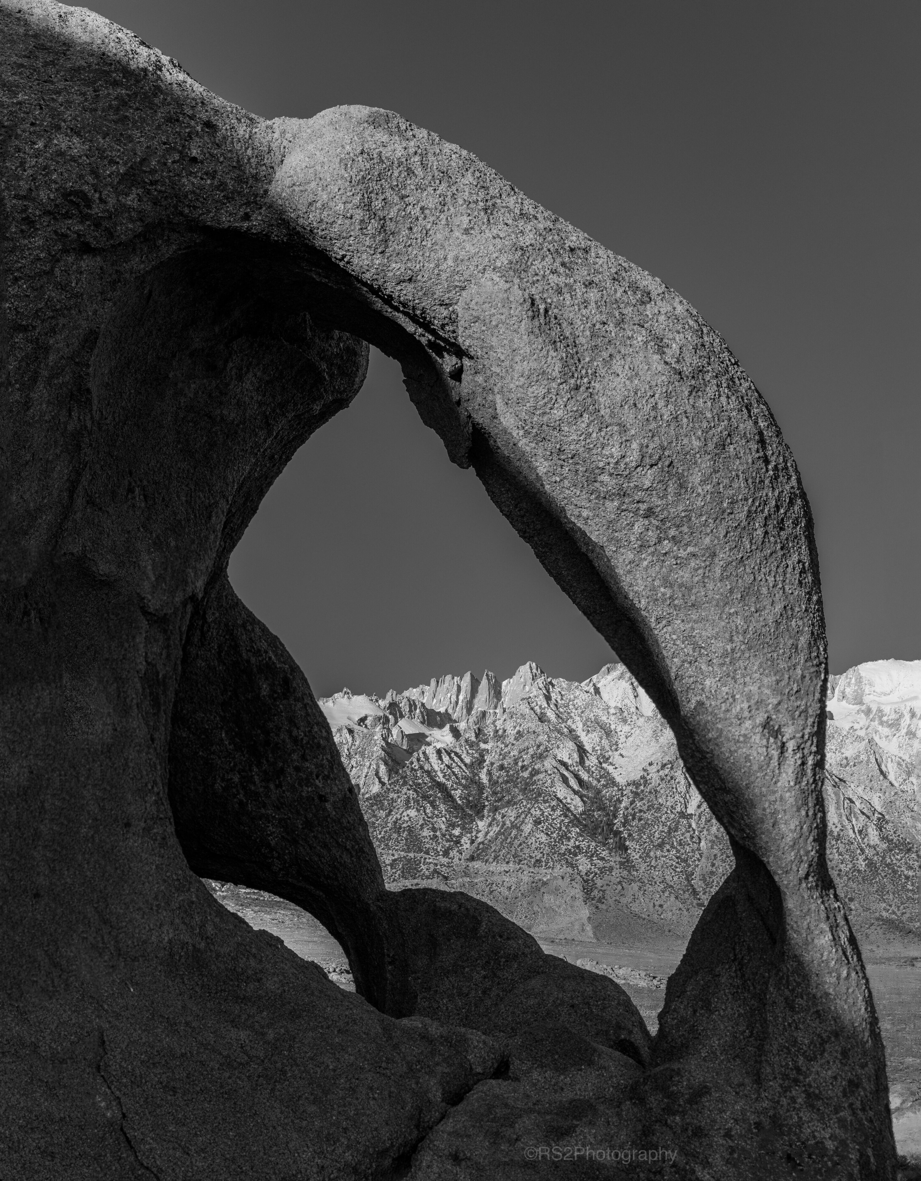 A large rock formation with a natural archway, rugged mountains in the background, and a clear sky.