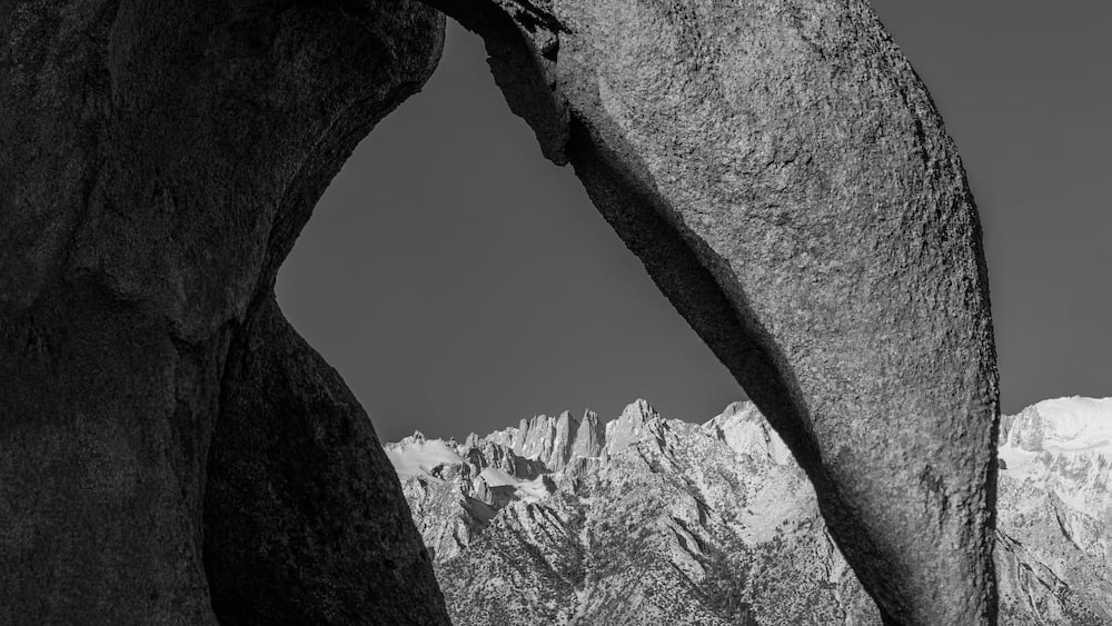 A large rock formation with a natural archway, rugged mountains in the background, and a clear sky.