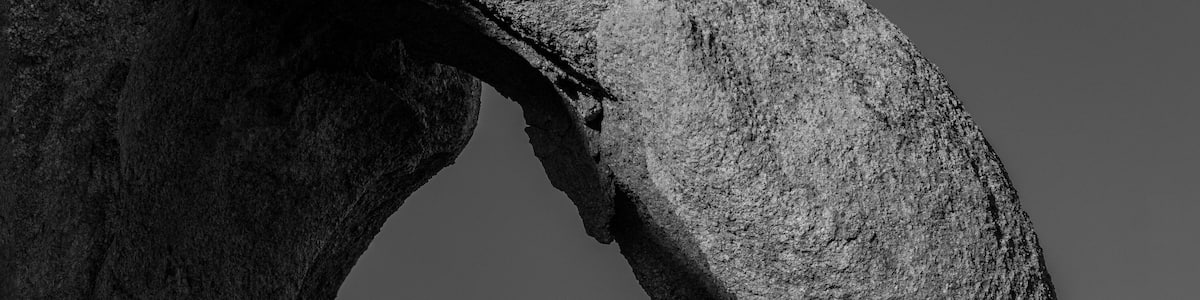 Une vaste formation rocheuse surmontée d’une arche naturelle, des montagnes accidentées en arrière-plan et un ciel dégagé.