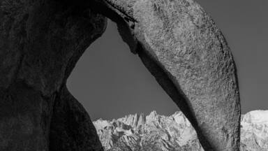 Uma grande formação rochosa com um arco natural, montanhas acidentadas ao fundo e céu claro.