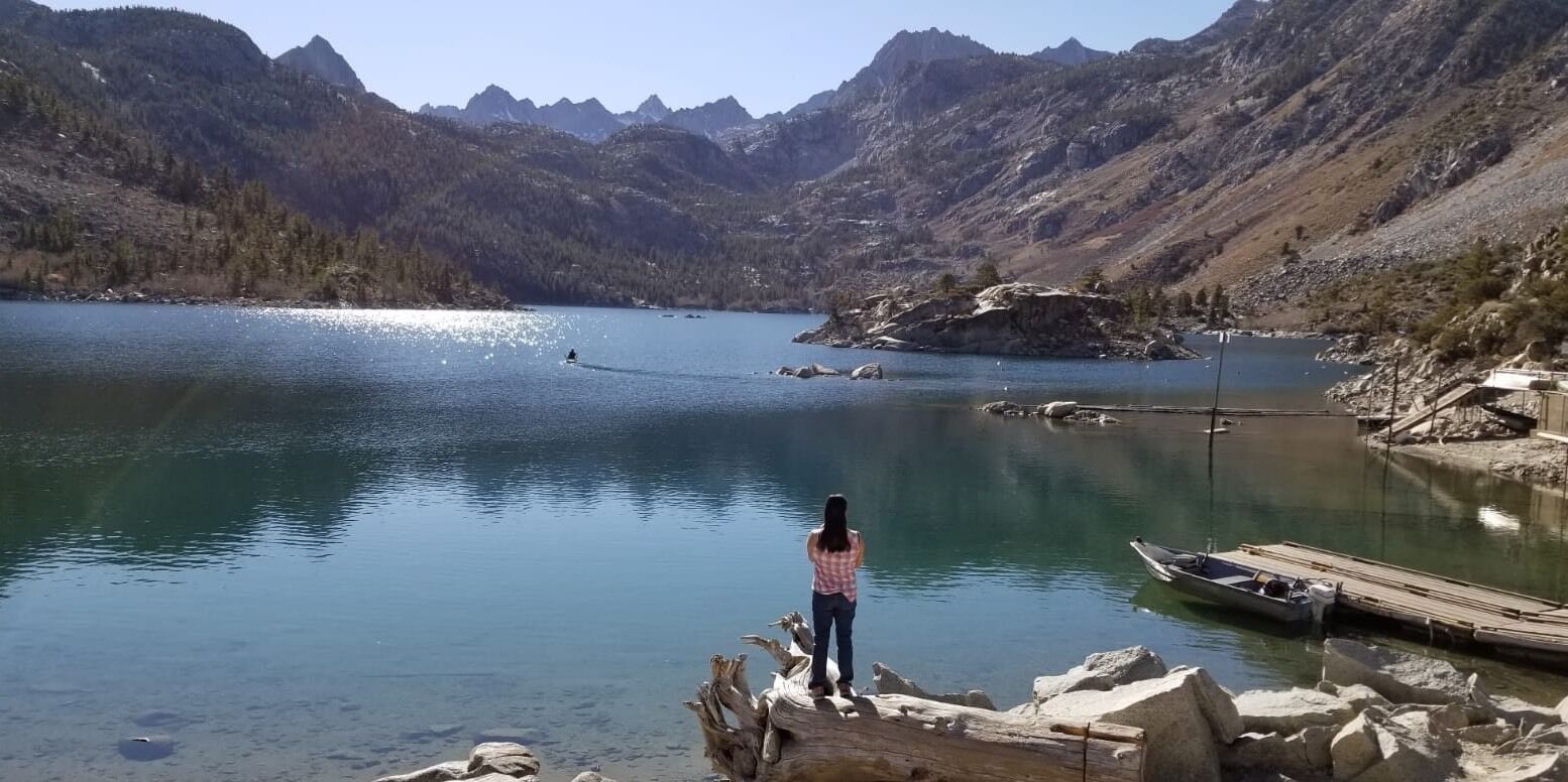 A person standing on a rocky outcrop overlooking a calm lake surrounded by mountains.