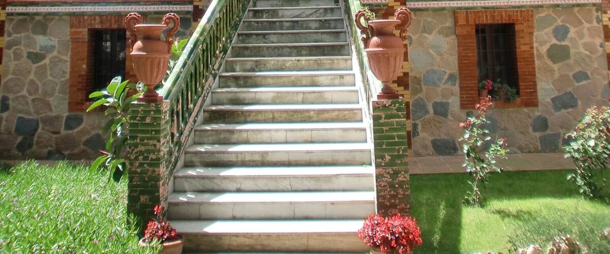 A large, ornate building with a grand staircase leading to a central door, surrounded by lush greenery.