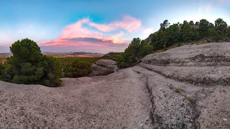 A rocky landscape with dense pine trees and a distant view of a valley under a colorful sky.