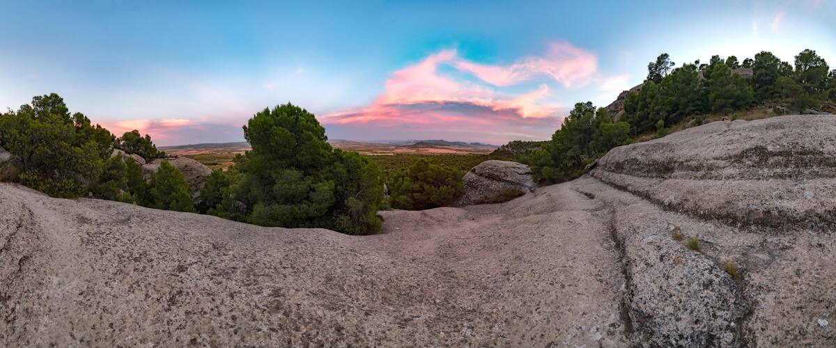 A rocky landscape with dense pine trees and a distant view of a valley under a colorful sky.