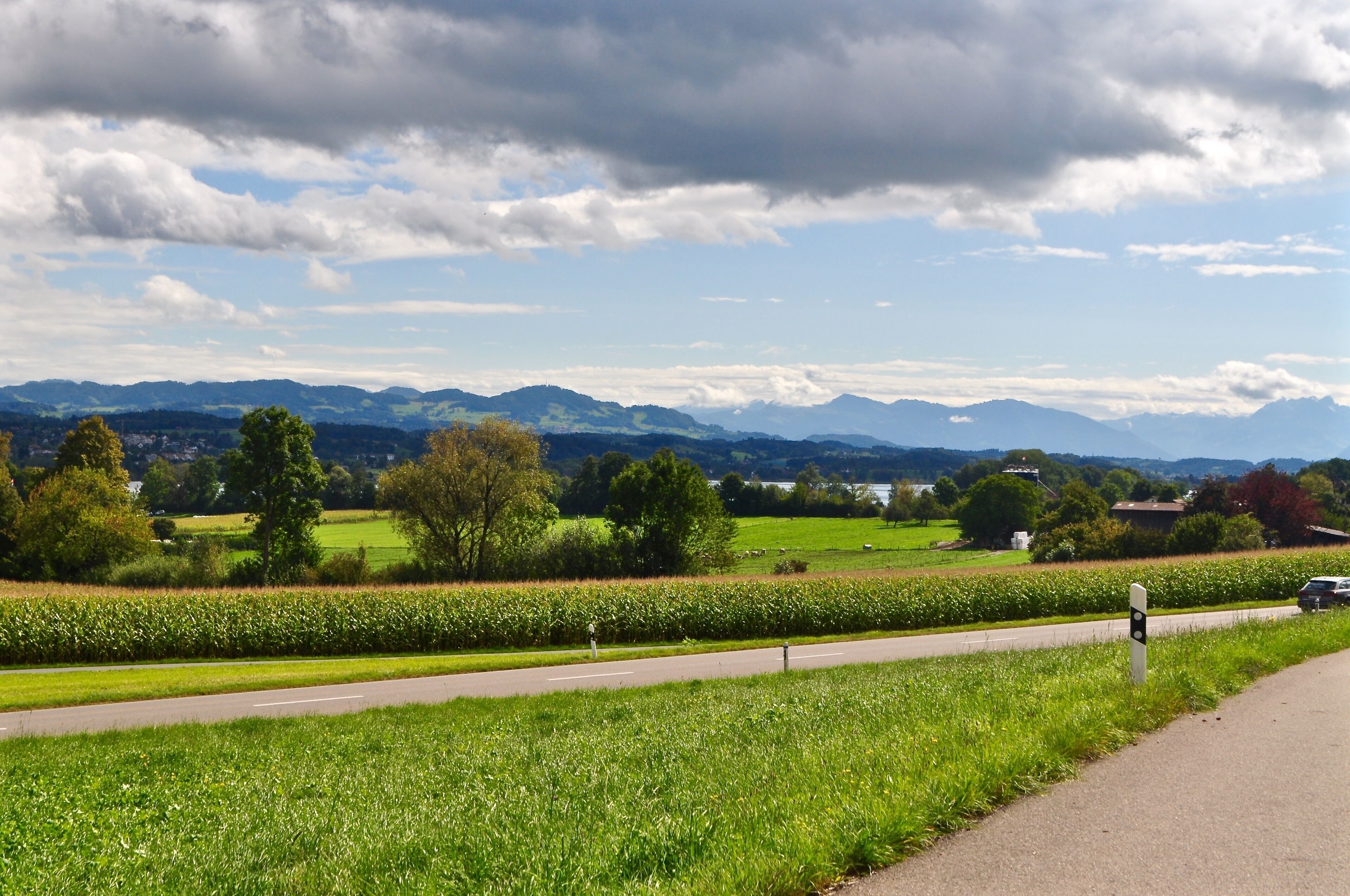 A rural landscape with a road, green fields, trees, and distant mountains under a partly cloudy sky.