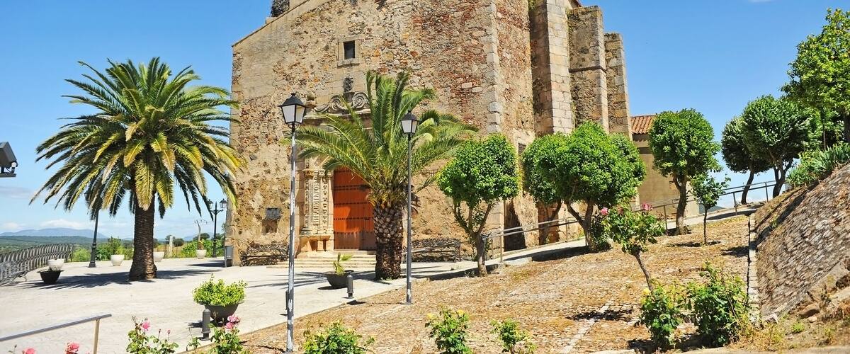 A historic stone building with a conical roof, palm trees, and a clear blue sky.