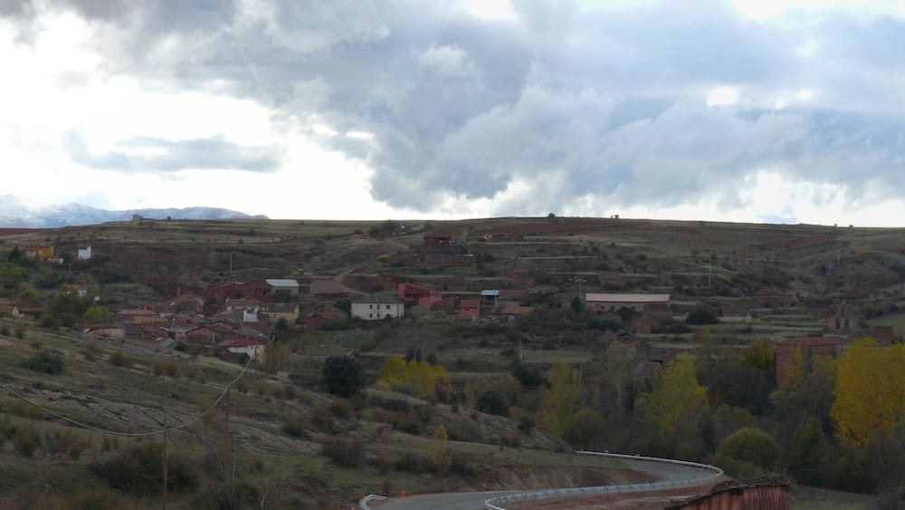 A rural landscape with a winding road, scattered houses, and a backdrop of mountains under a cloudy sky.