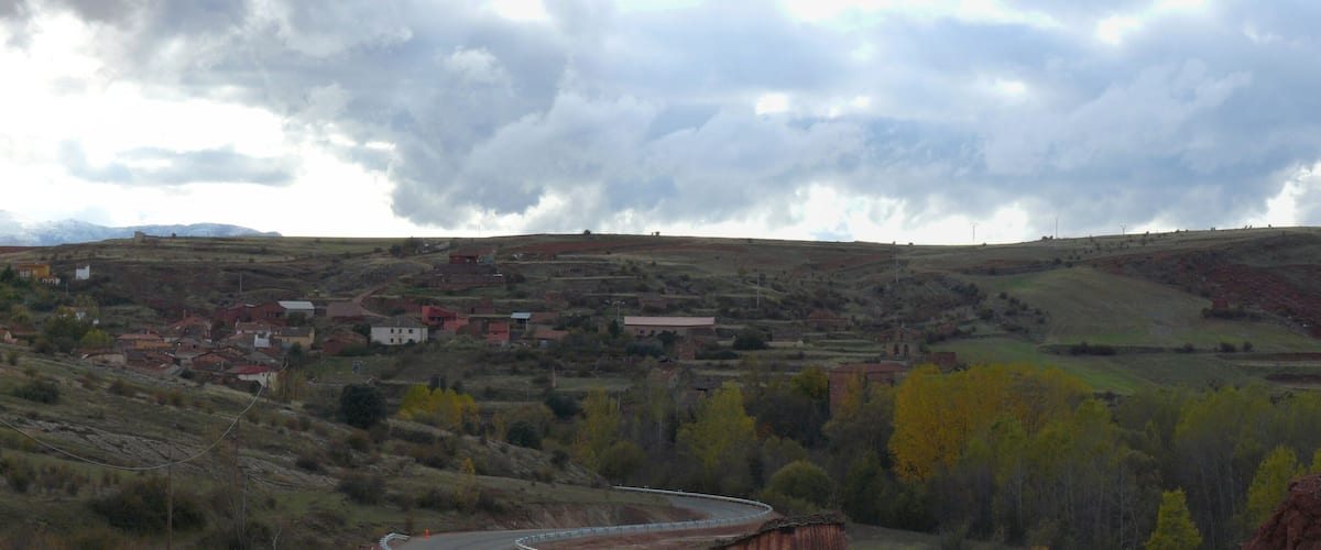 A rural landscape with a winding road, scattered houses, and a backdrop of mountains under a cloudy sky.