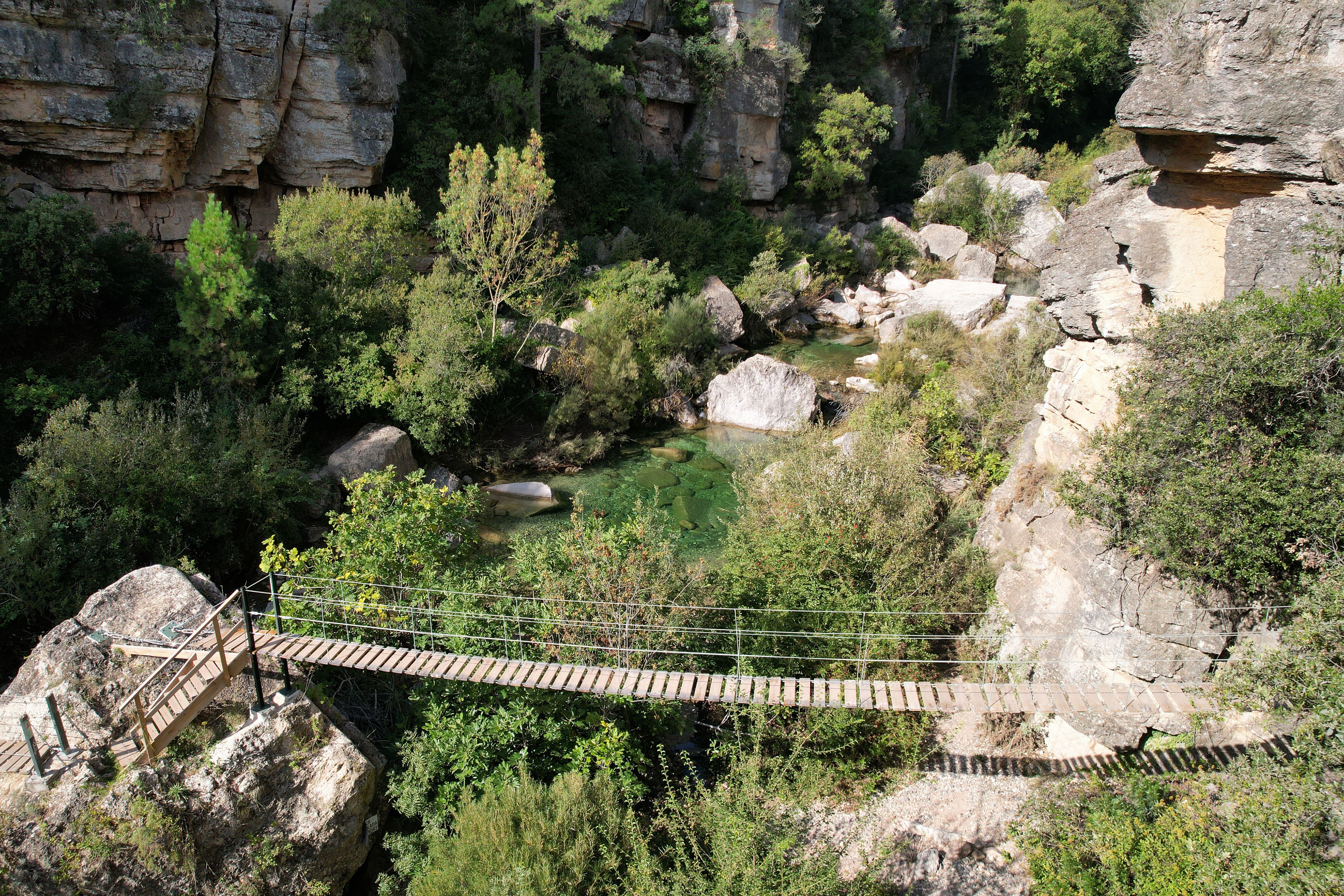 Un ponte sospeso in legno che attraversa un torrente cristallino, circondato da scogliere rocciose e rigogliosa vegetazione.