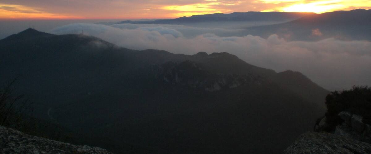 A mountain range with a sunrise or sunset, clouds below the peaks, and a clear sky.