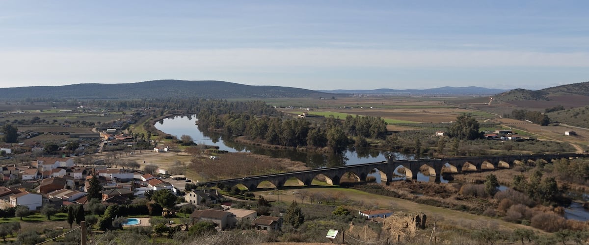 A river flowing through a valley with a bridge, surrounded by hills and a small town.
