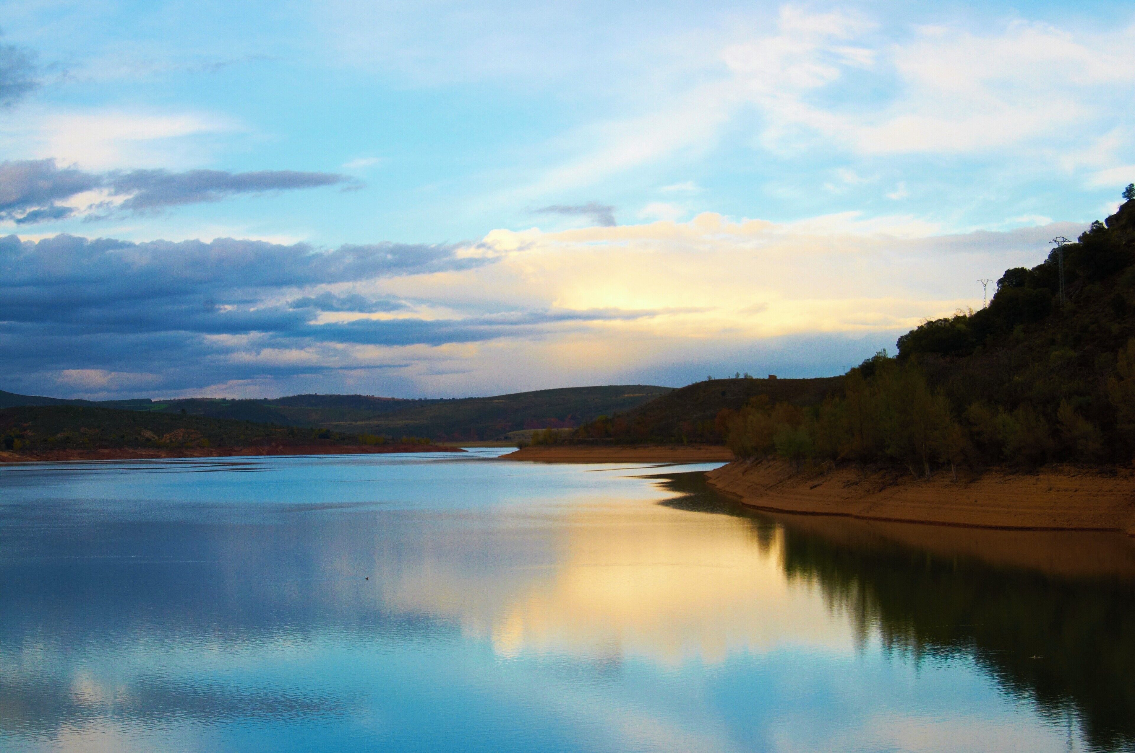 A calm river reflecting the sky, surrounded by hills and sparse trees.