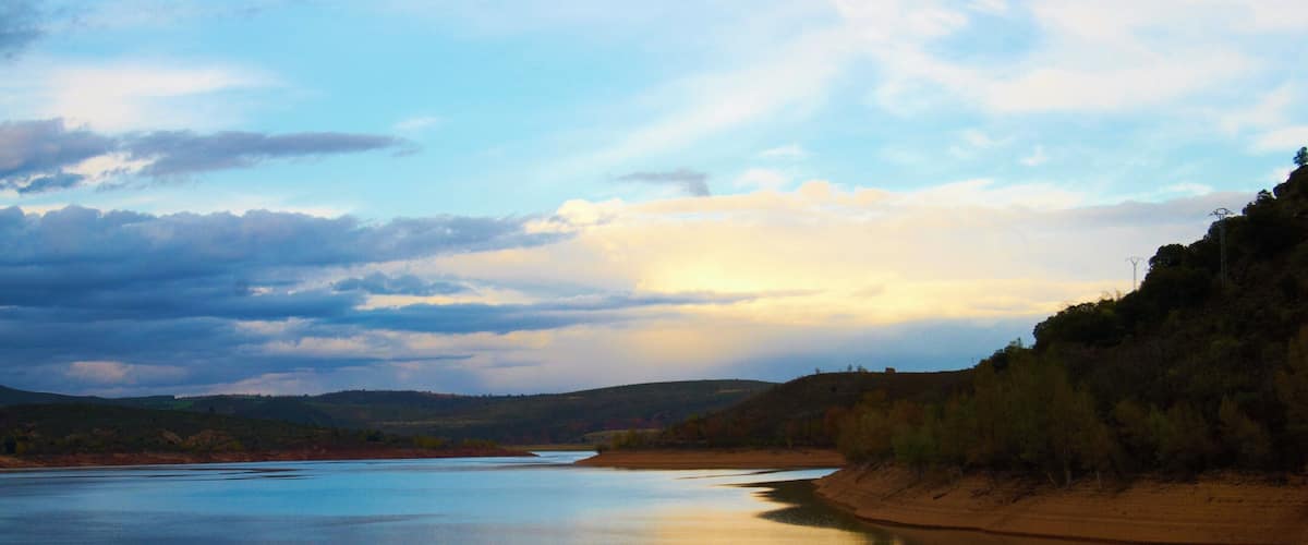A calm river reflecting the sky, surrounded by hills and sparse trees.