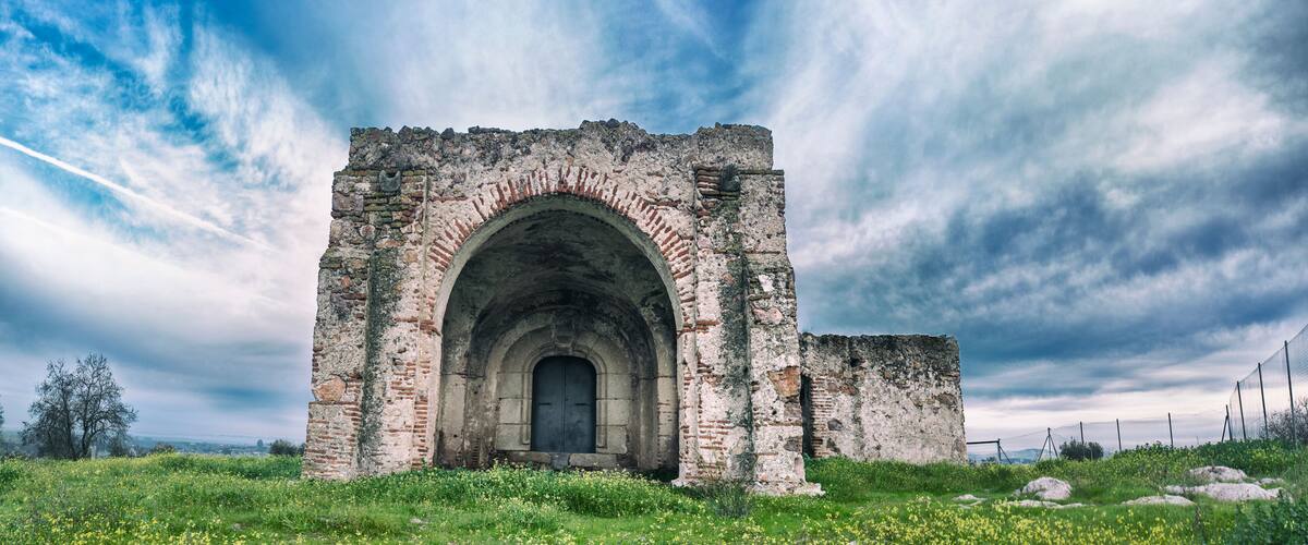 An ancient stone archway with a dark door, surrounded by lush greenery and a cloudy sky.
