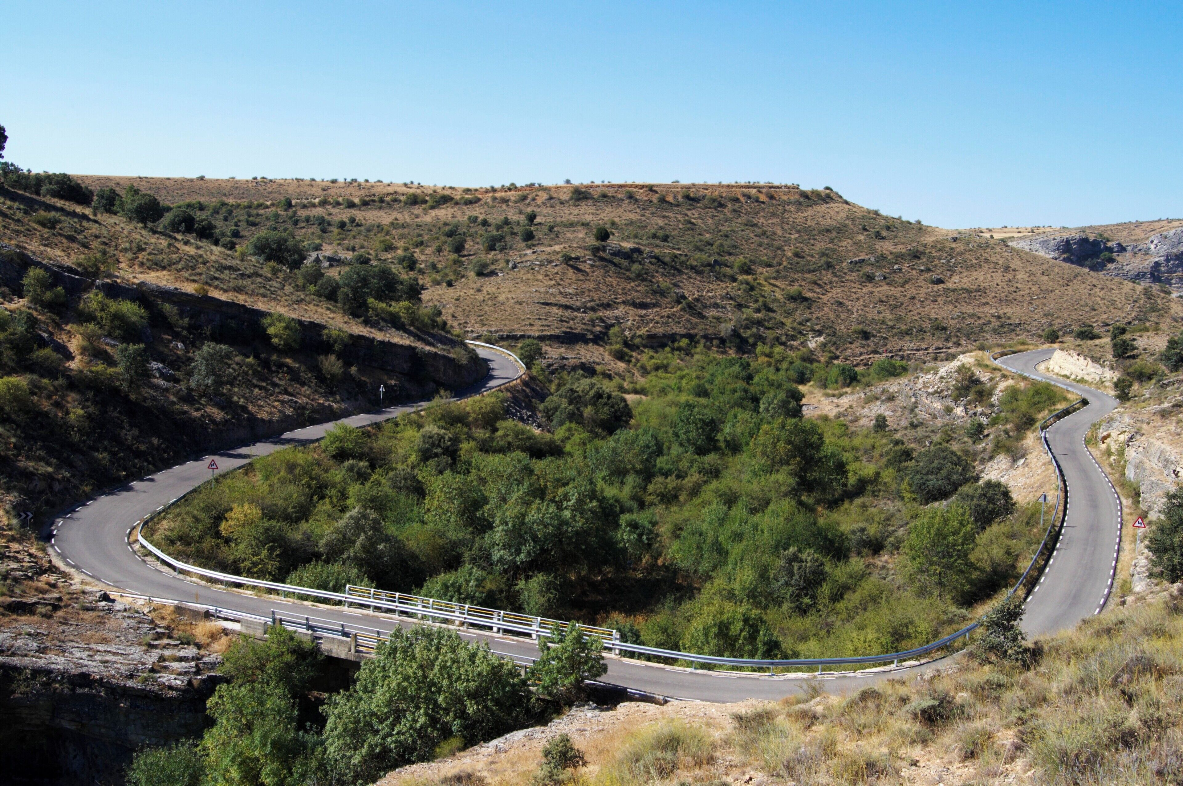 A winding mountain road with guardrails, surrounded by dry vegetation and rocky terrain.