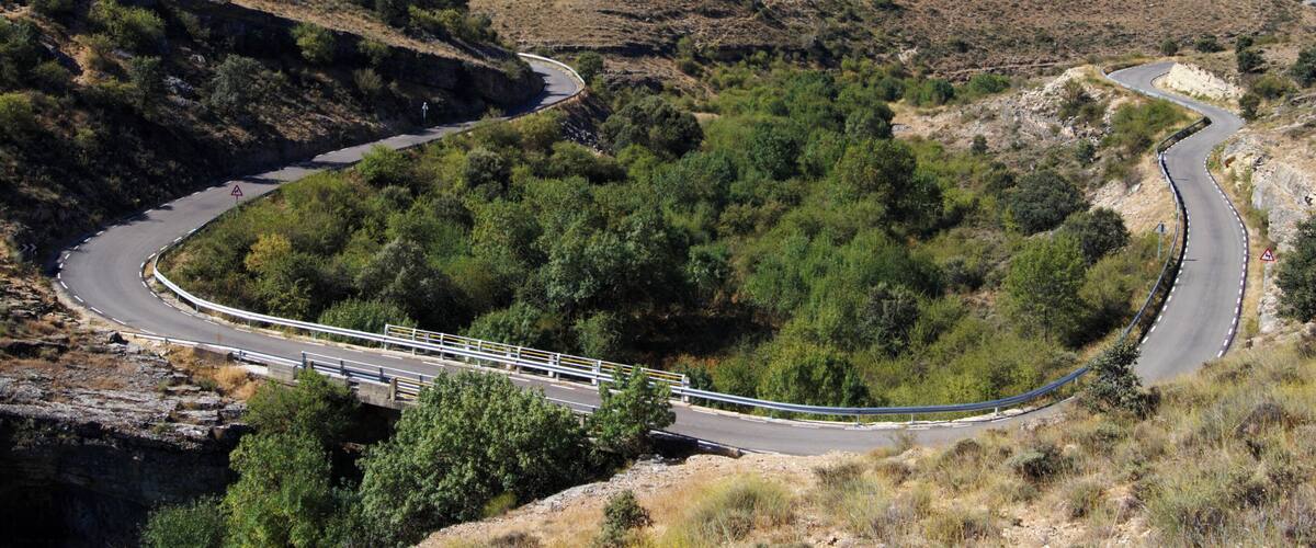 A winding mountain road with guardrails, surrounded by dry vegetation and rocky terrain.