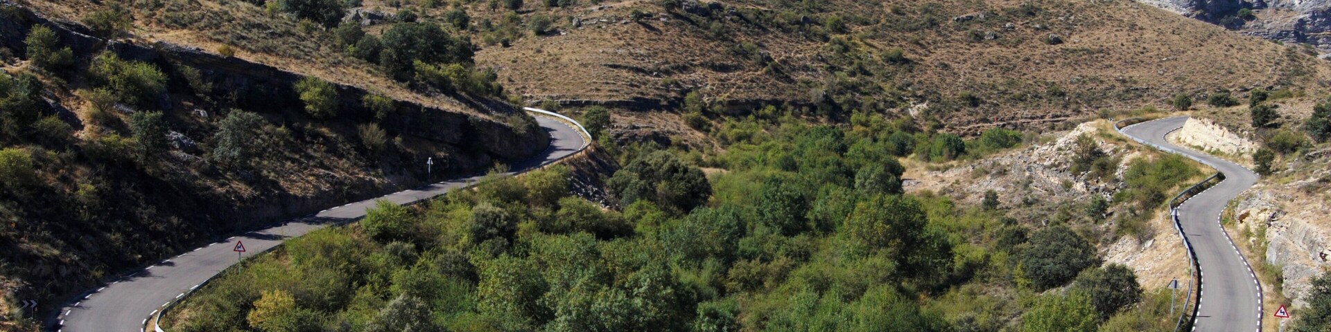 A winding mountain road with guardrails, surrounded by dry vegetation and rocky terrain.