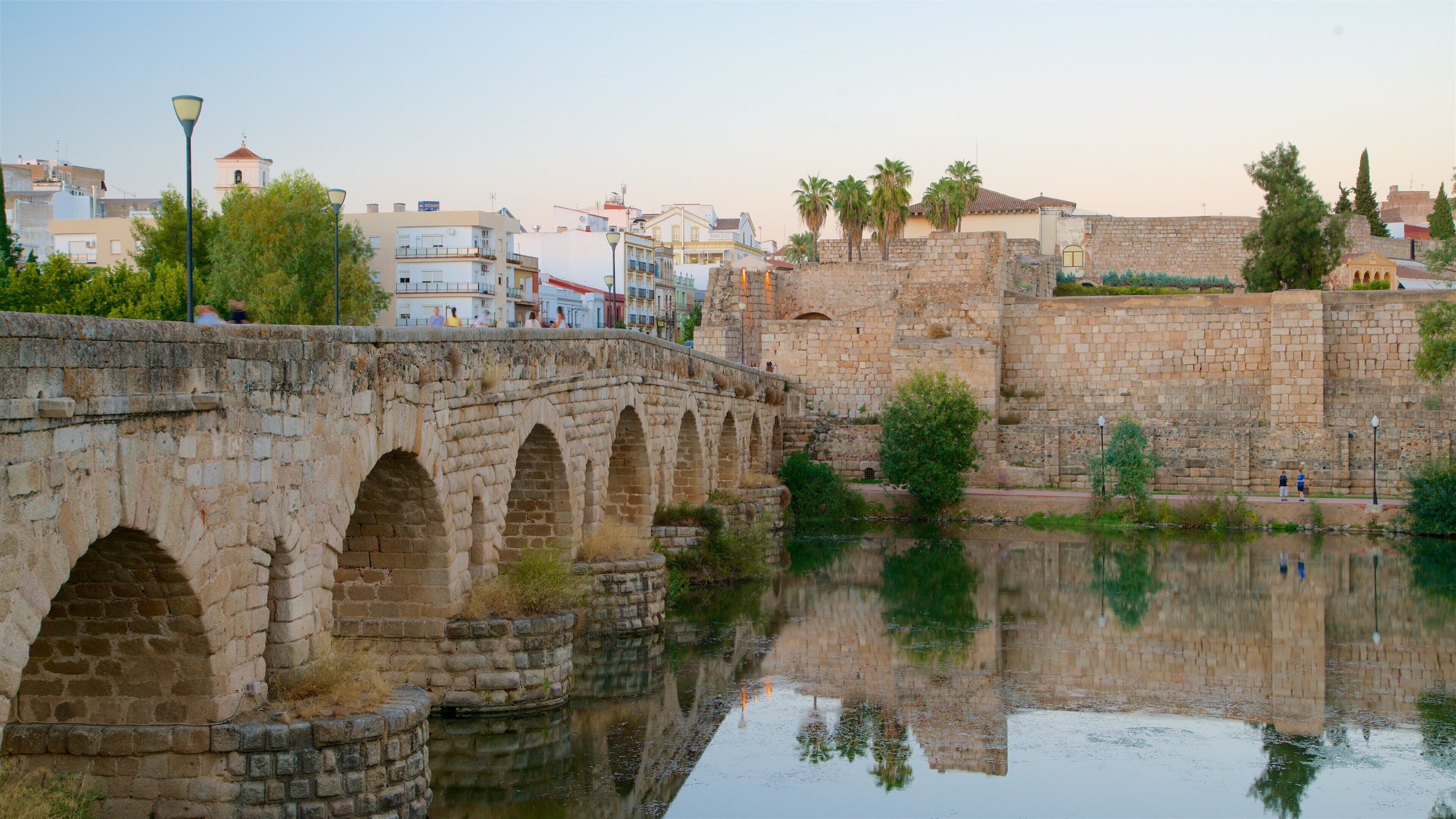 Un puente de piedra sobre un río tranquilo, con edificios y un muro al fondo.