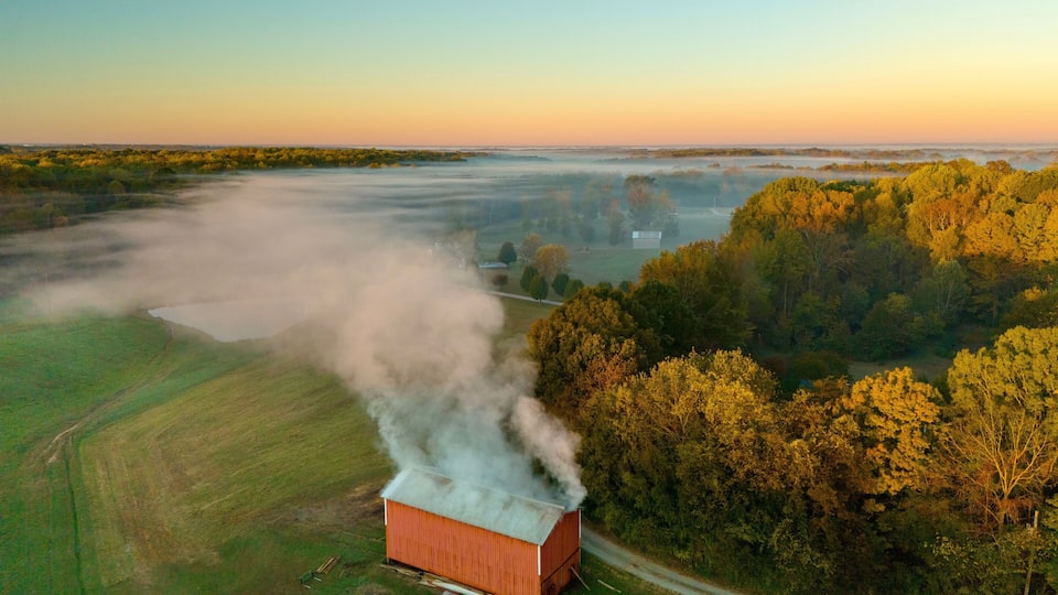 An aerial view of a red barn with smoke, surrounded by green fields and trees.