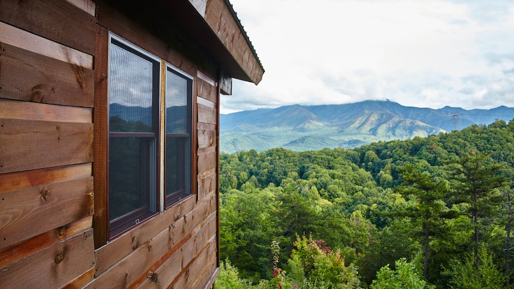 A wooden cabin with a view of a dense forest and mountains in the background.