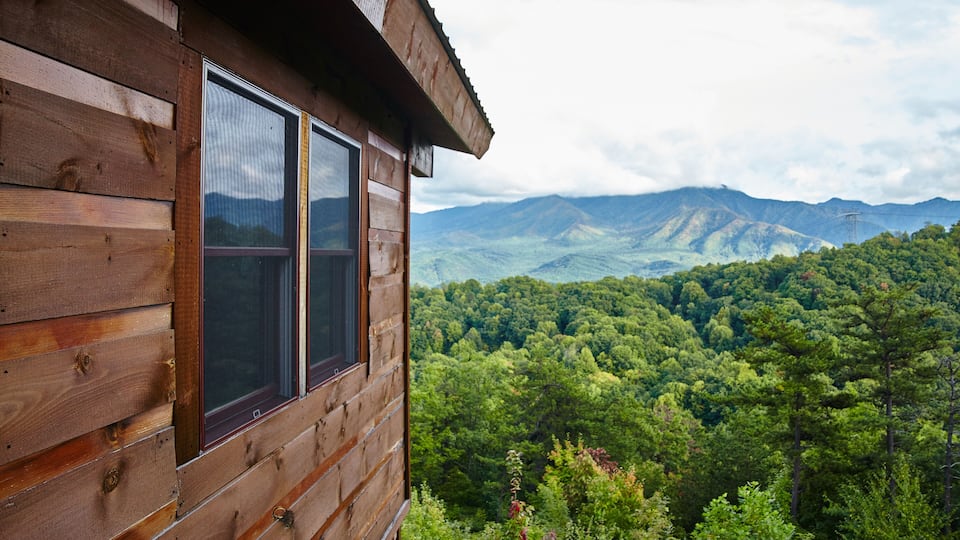A wooden cabin with a view of a dense forest and mountains in the background.