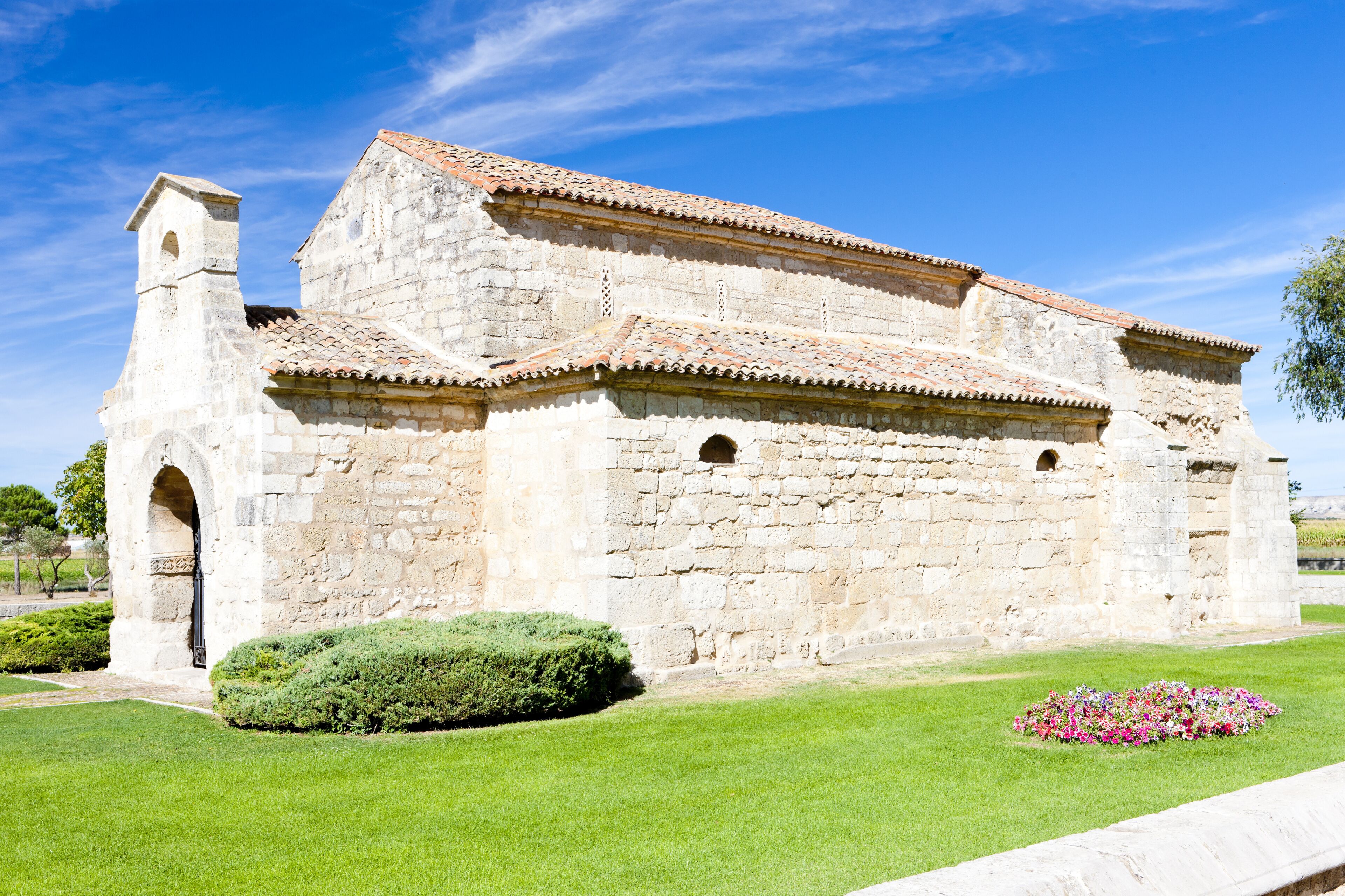 A historic stone church with a bell tower and tiled roof, surrounded by well-maintained grass and flower beds.