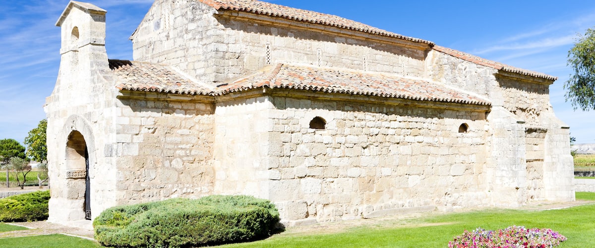A historic stone church with a bell tower and tiled roof, surrounded by well-maintained grass and flower beds.