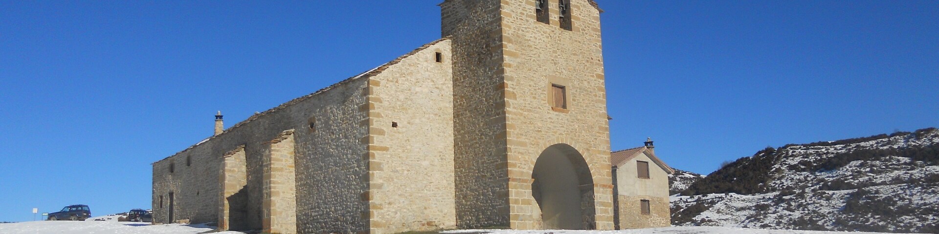 A stone church with a bell tower and arched doorway, surrounded by snow-covered ground and a clear blue sky.