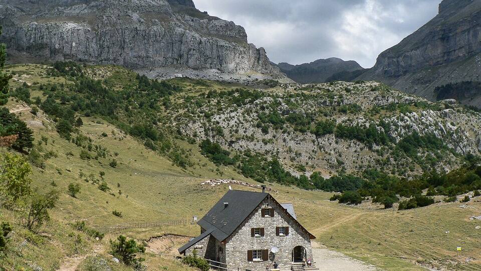A stone house with a sloped roof in a mountainous area with green vegetation and rocky cliffs.