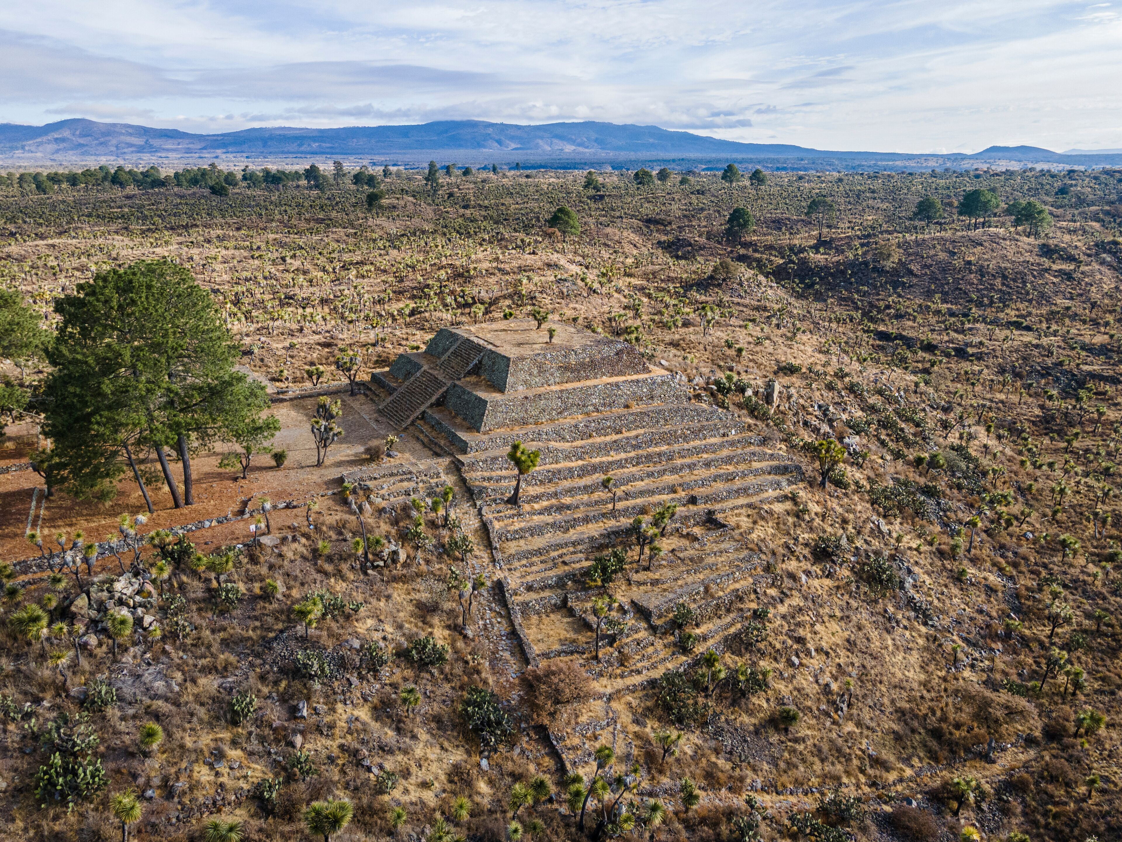 A large, ancient pyramid complex in a dry, arid landscape with sparse vegetation and distant mountains.