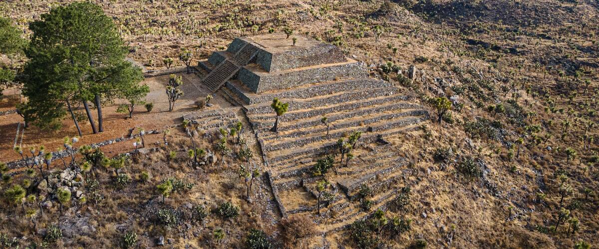 A large, ancient pyramid complex in a dry, arid landscape with sparse vegetation and distant mountains.