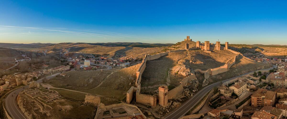 A historic fortress with multiple towers, a winding road, and a town in the valley below.
